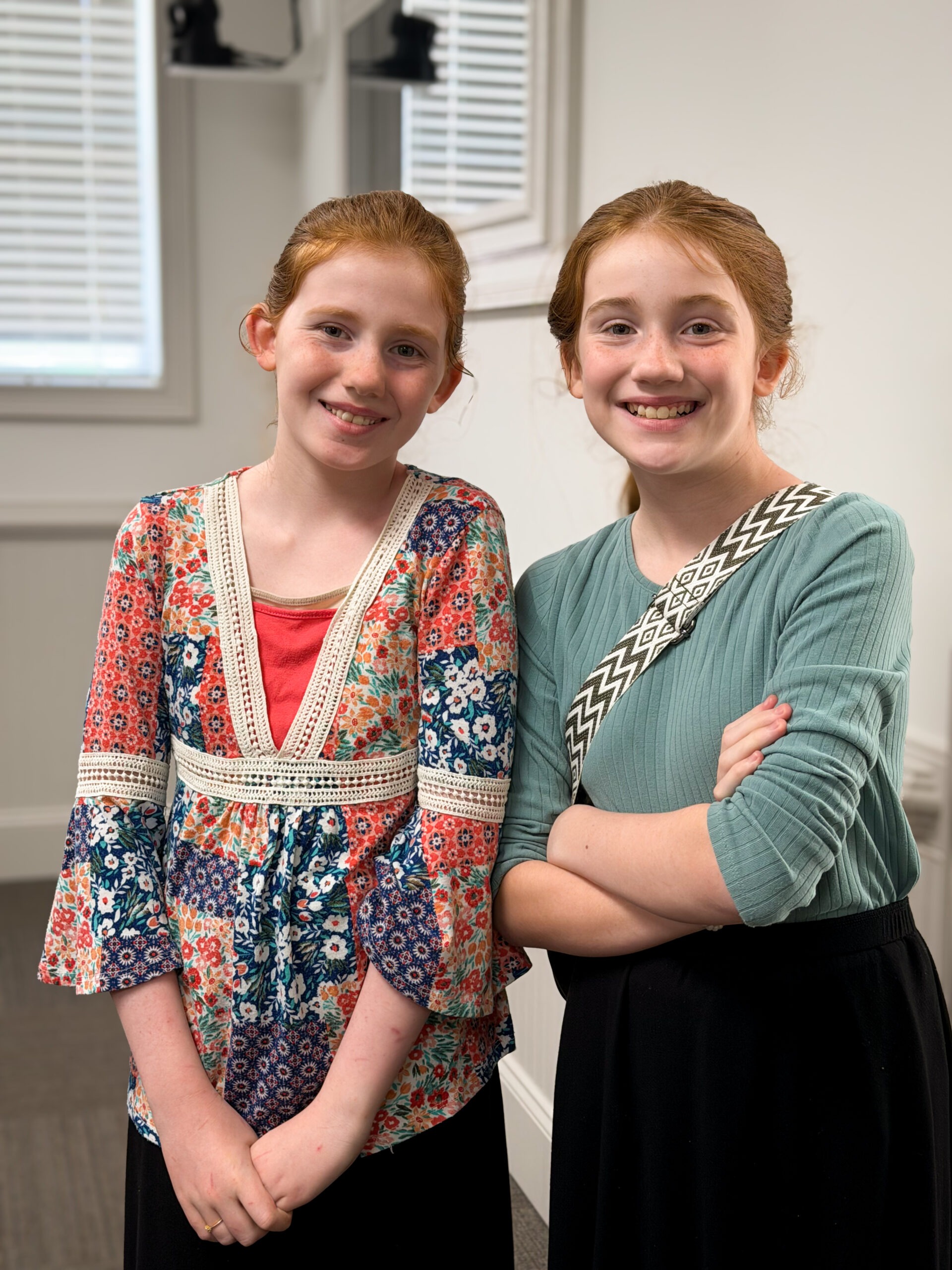 Two smiling girls stand side by side in a bright room. One wears a colorful floral blouse, while the other is dressed in a teal top.