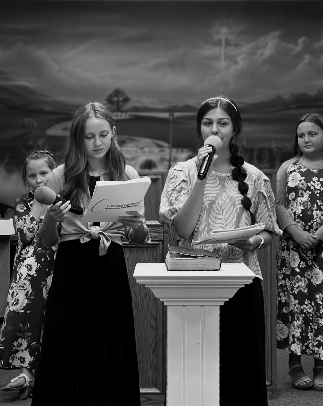 Two young women speak at a podium in a church, one holding a microphone and the other reading from a script. Several girls listen attentively in the background.