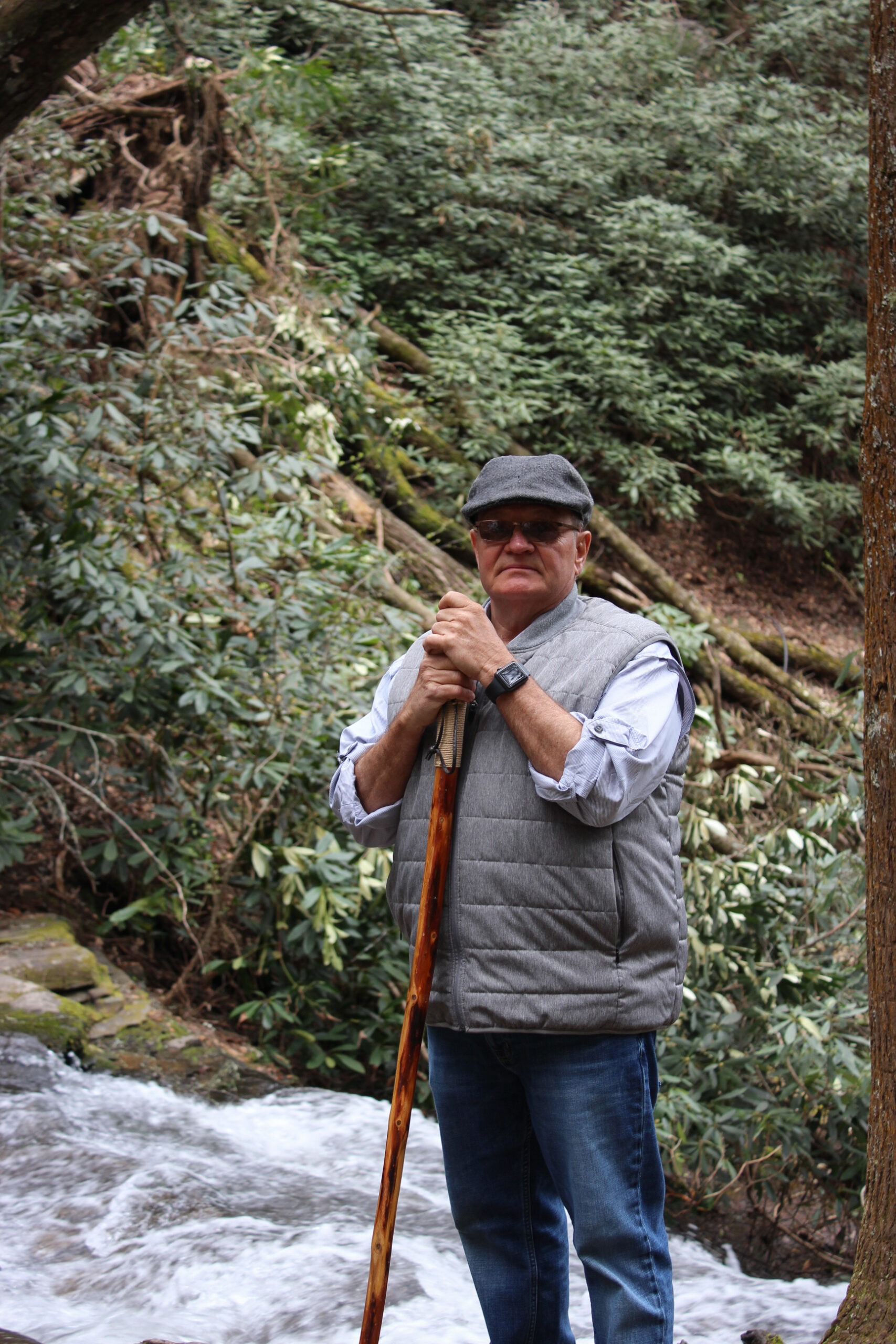 Man standing by a flowing stream in a forest, wearing sunglasses, a gray vest, and a flat cap, holding a wooden cane.