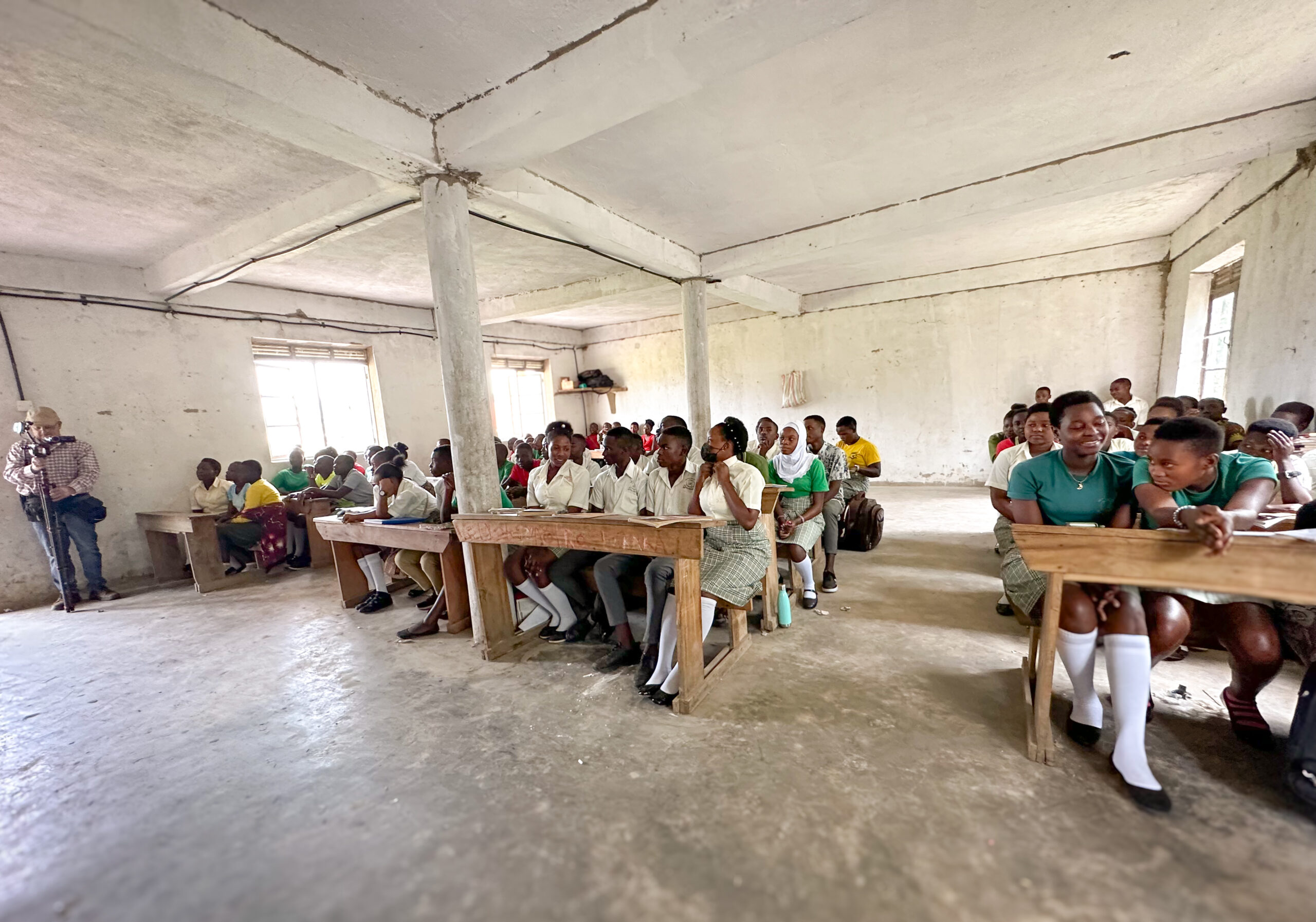 A classroom filled with students attentively listening, seated at wooden benches. The educational atmosphere highlights engagement in learning.
