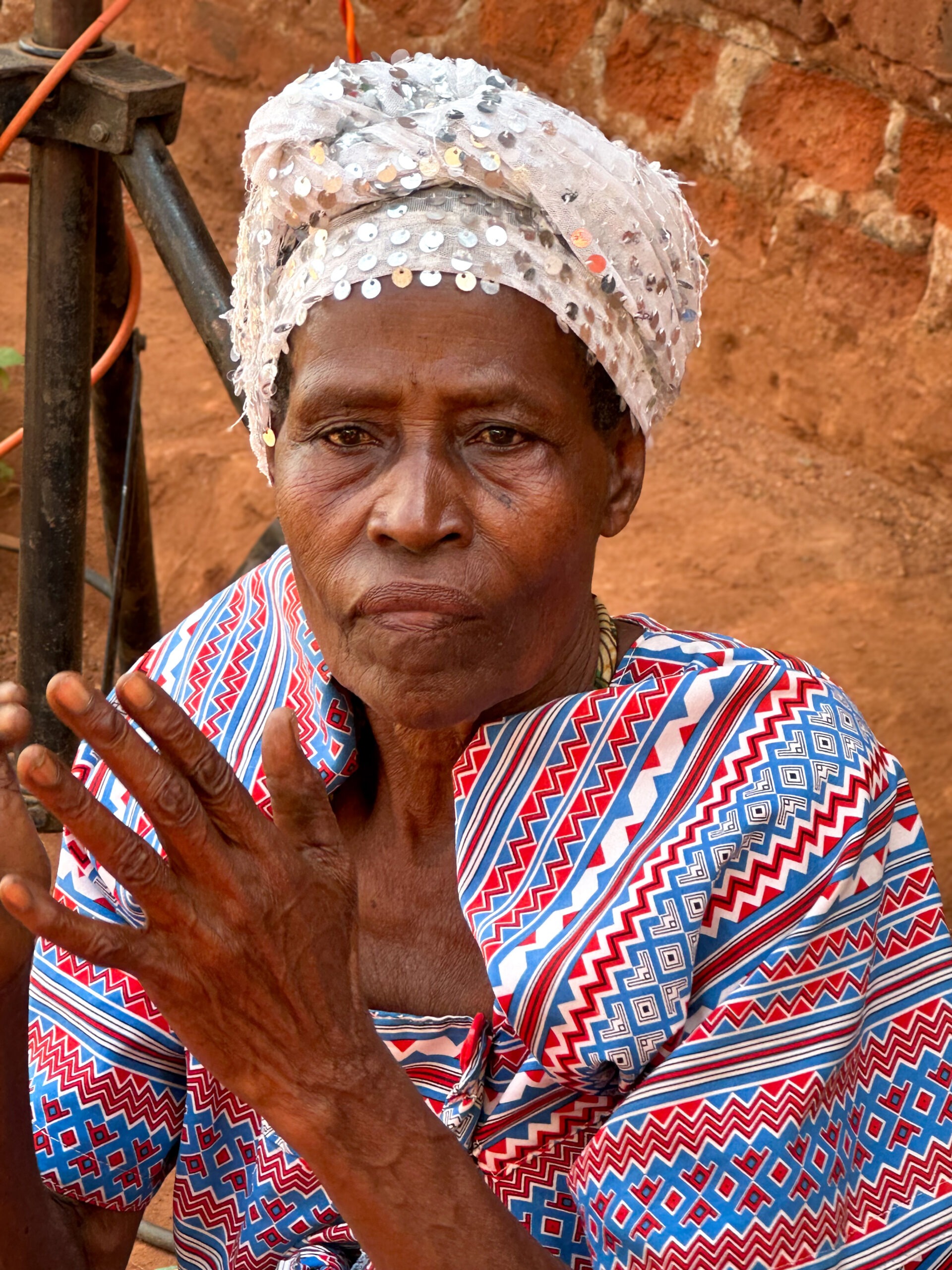 Elderly woman with a patterned red and blue garment, wearing a sparkly white headscarf, sits against a rustic brick wall, looking contemplative.