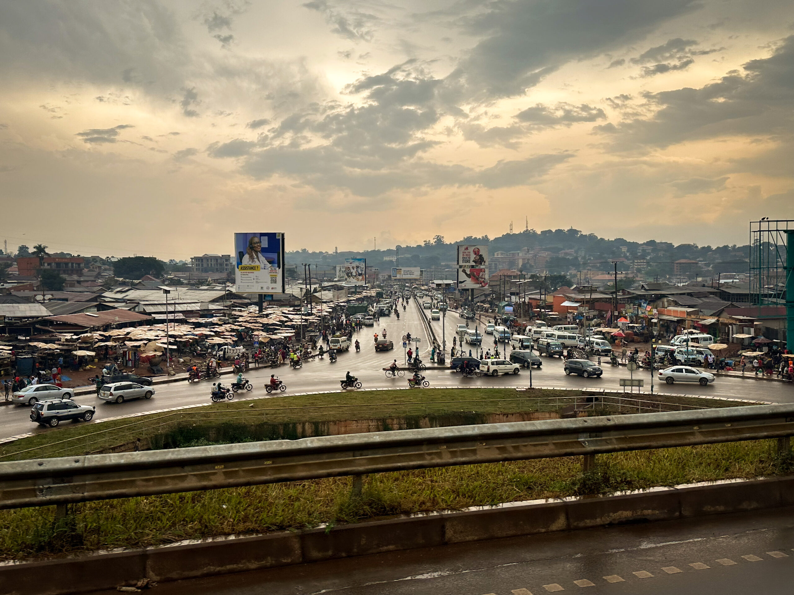 Busy urban road scene showing a marketplace with vendors and vehicles. A large billboard is visible, indicating commercial activity in the area.