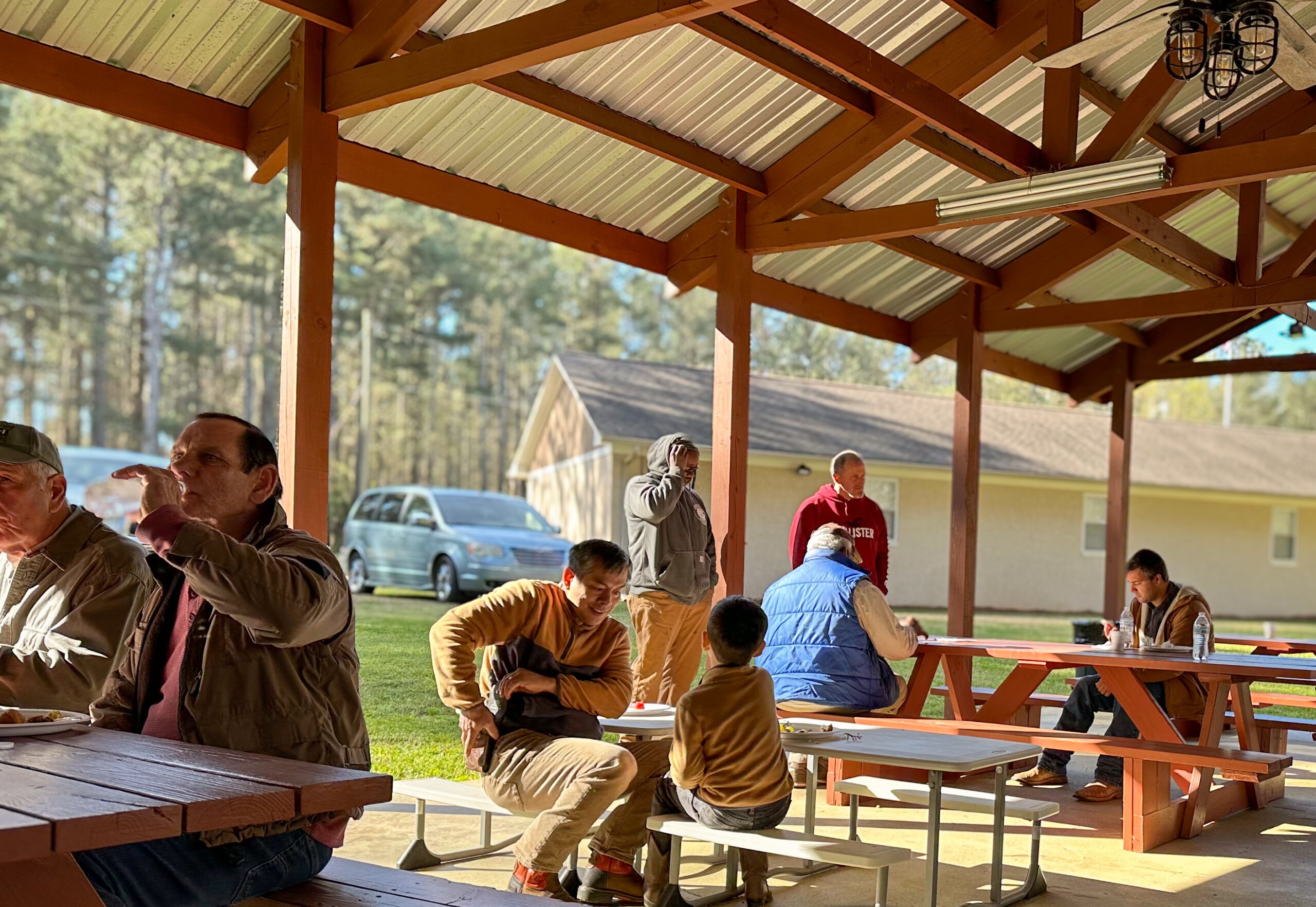 A diverse group of people sharing a meal under a shelter, with trees in the background. The setting emphasizes community and fellowship.