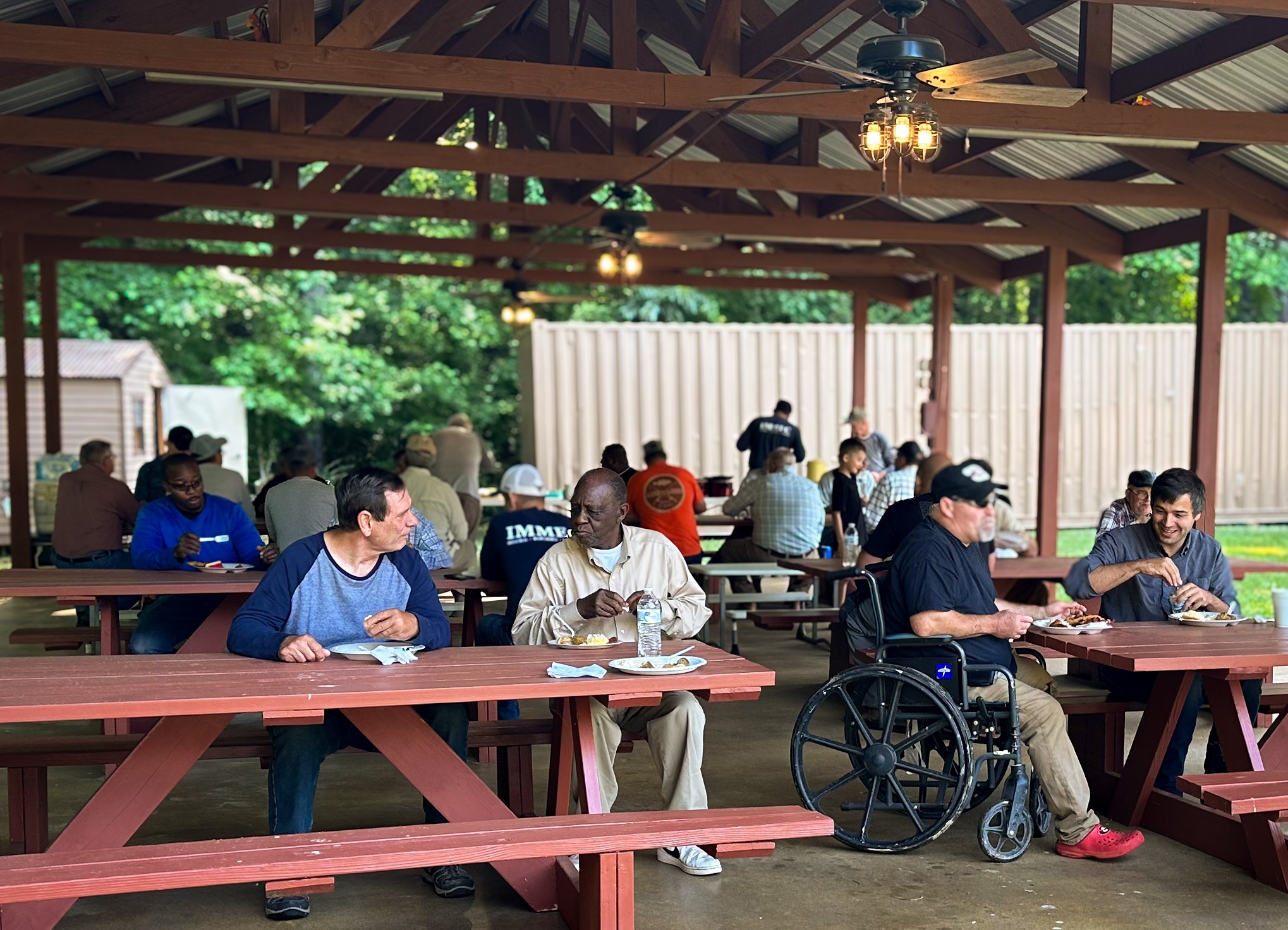 Community members gather for a meal under a pavilion, sharing food and conversation, highlighting social connections and togetherness.