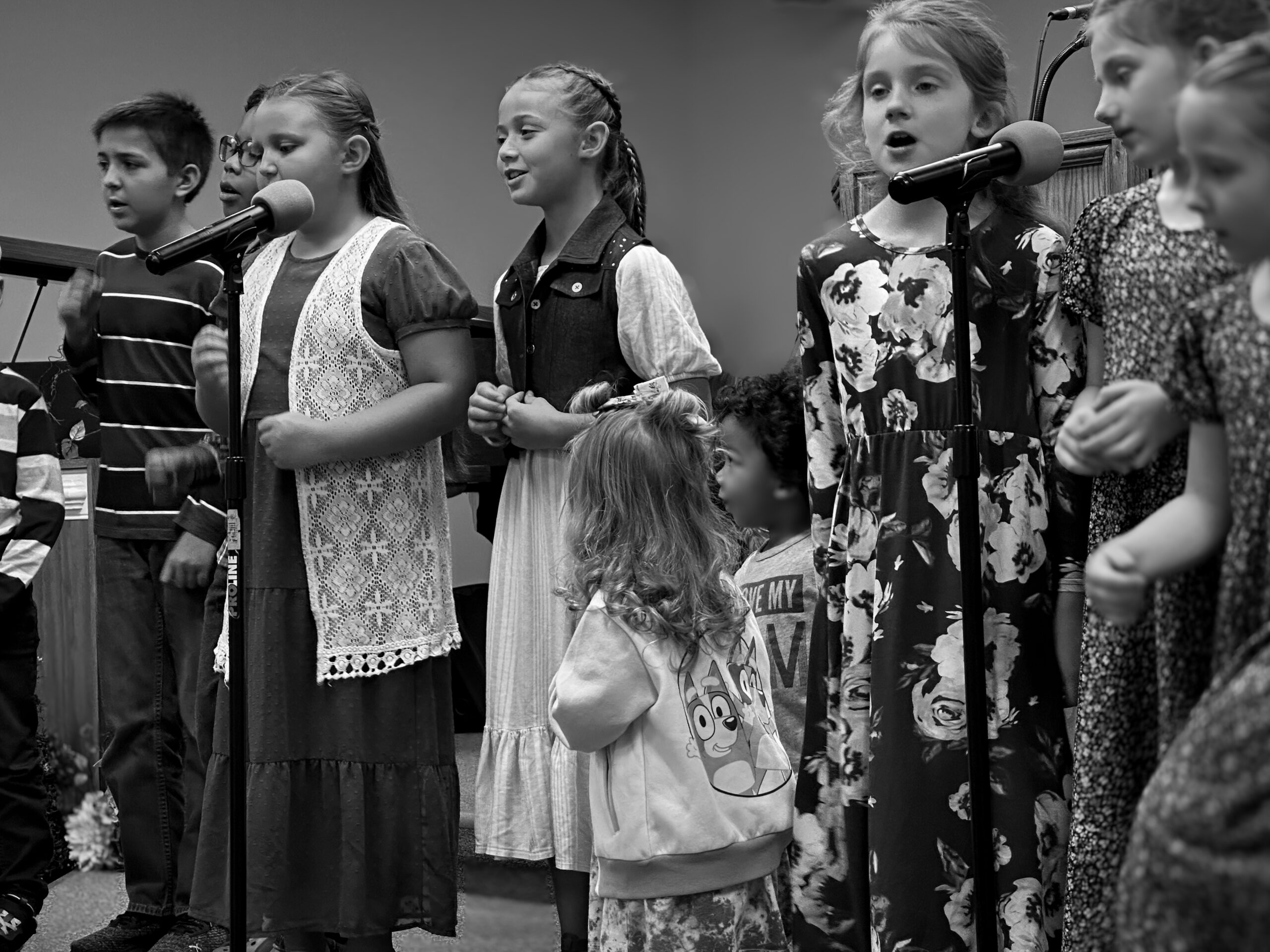 Children perform together at a microphone, expressing joy through song. The black-and-white image highlights their diverse outfits and engagement.