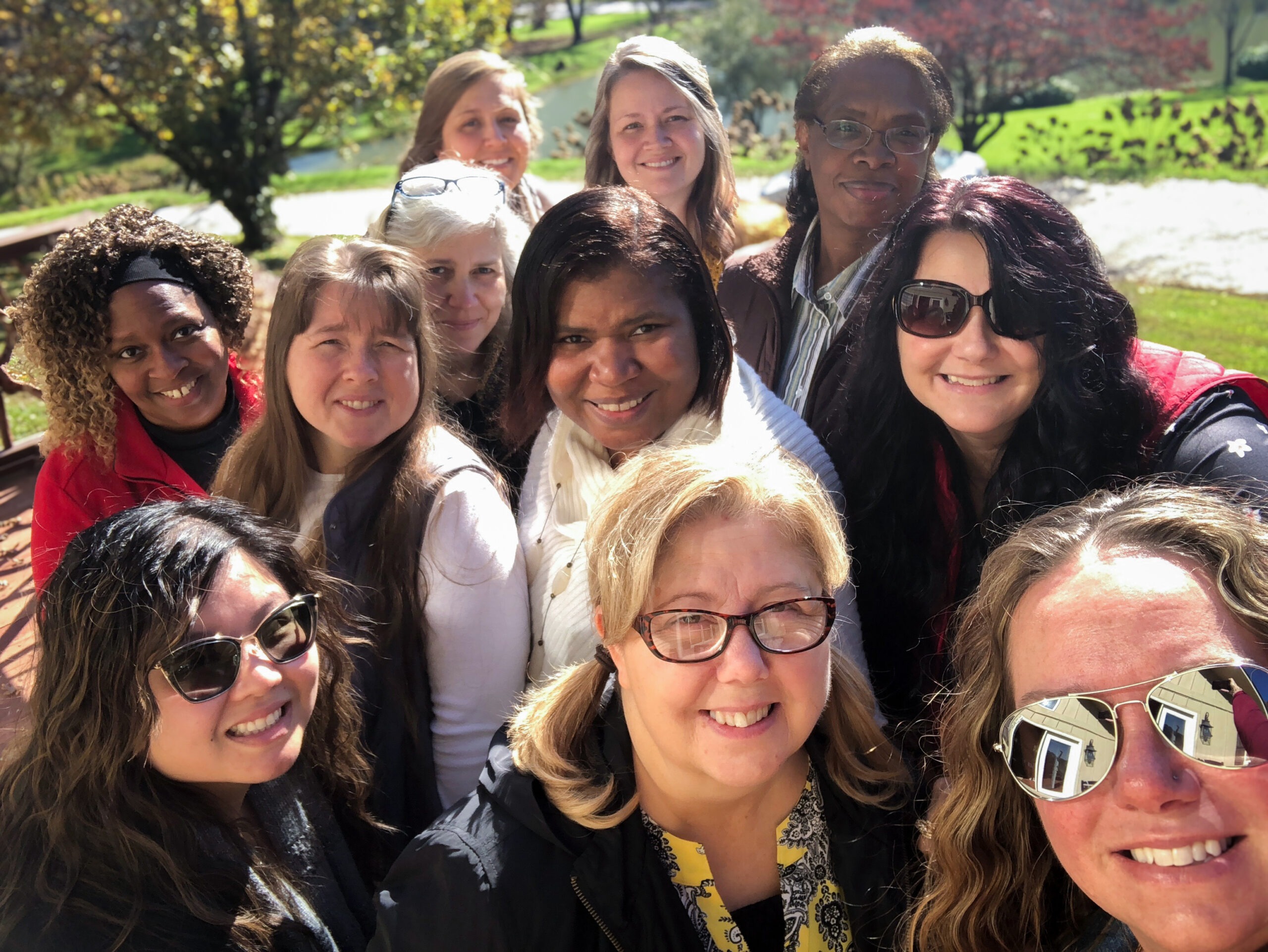 A group of twelve women smiles for a photo outdoors, surrounded by autumn foliage and greenery, capturing a moment of camaraderie.