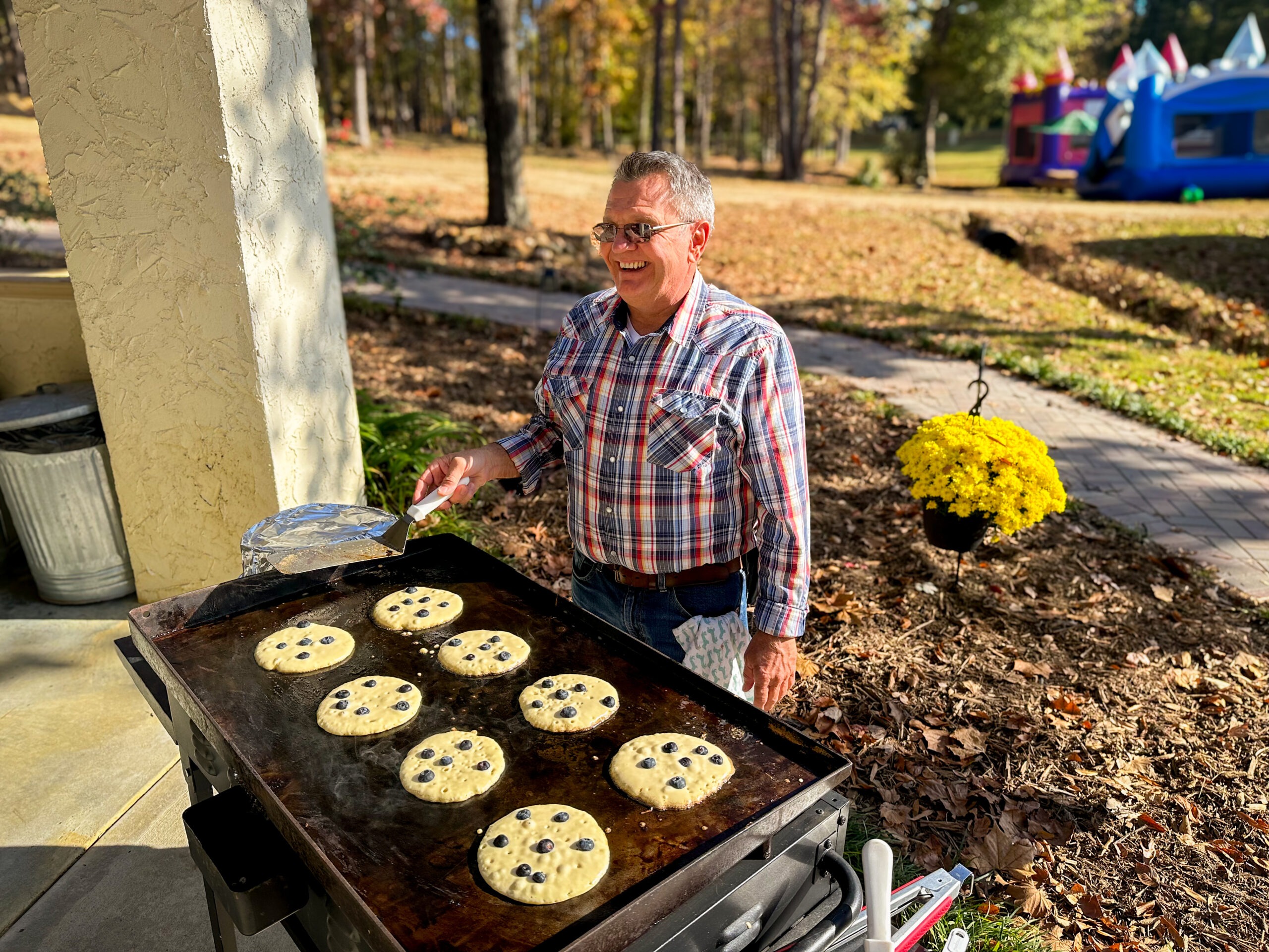 A smiling man in a plaid shirt cooks blueberry pancakes on a griddle outdoors, with autumn foliage and a bounce house in the background.